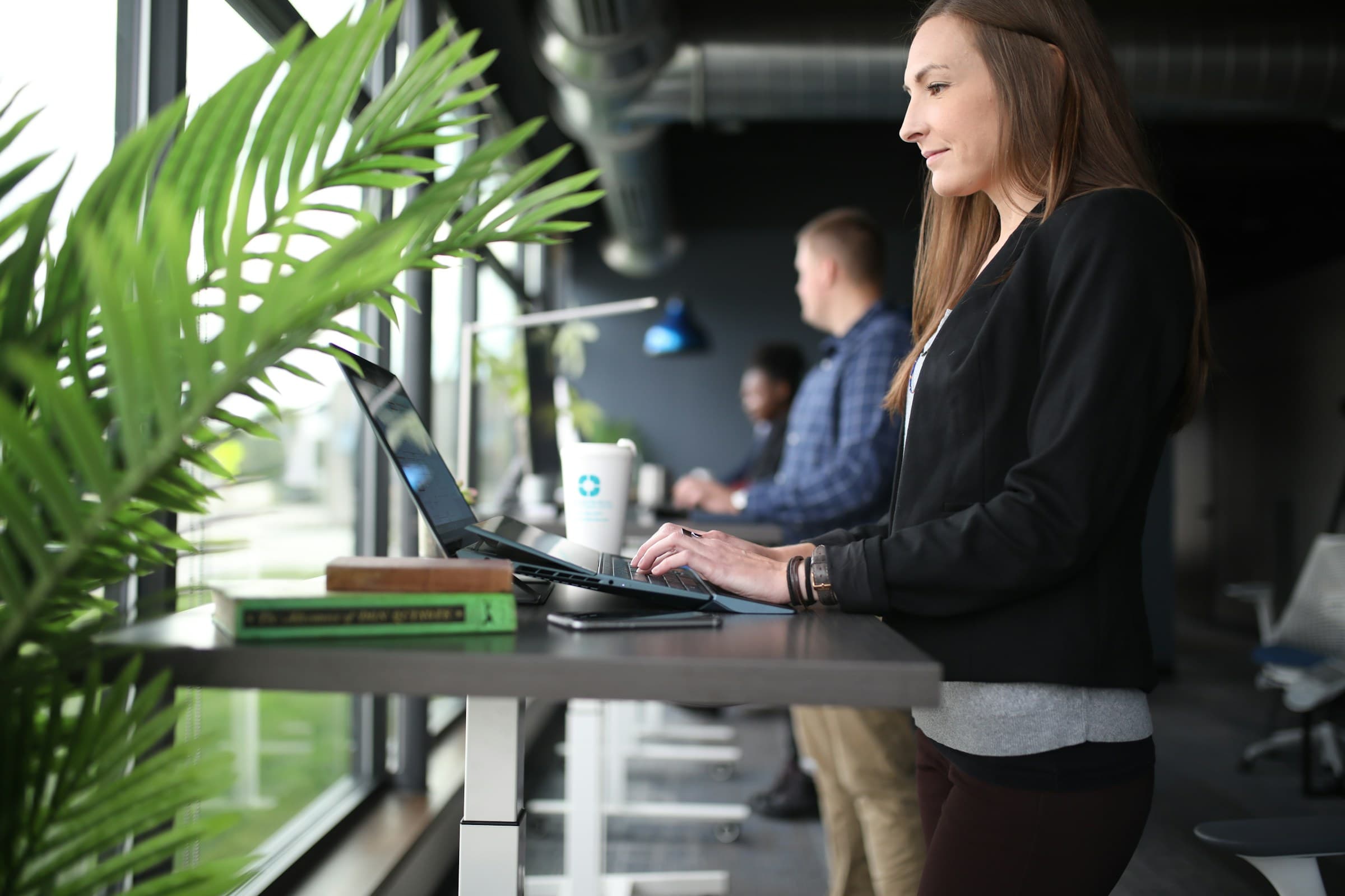 A woman at a standing desk by a wide window with plants