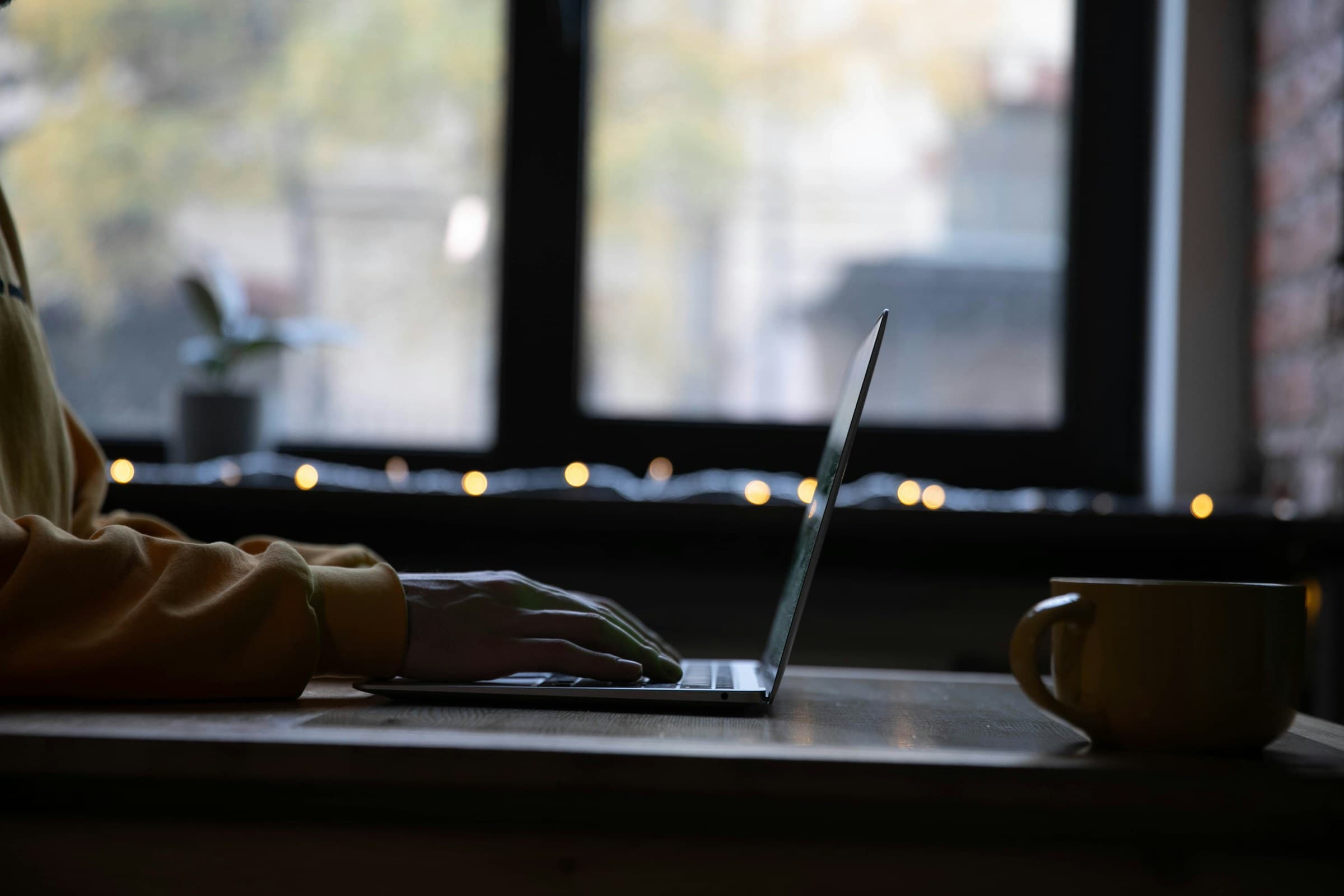 A laptop by a window with string lights and a mug